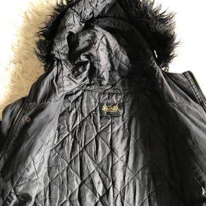May include: A black jacket with a quilted interior lining and a hood featuring black faux fur trim. The jacket has a central zipper closure and a black label with the gold text Von Dutch stitched into the inner collar area of the garment.