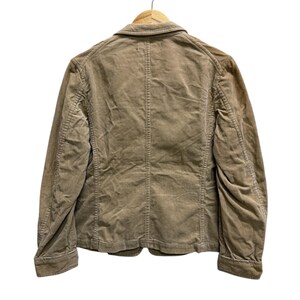 Puede incluir: Chaqueta de pana color beige, colgada de una percha de madera. La chaqueta presenta cierre de botones, cuello cl&aacute;sico y mangas largas. La vista trasera revela costuras verticales y un dise&ntilde;o estructurado, ideal para uso diario.