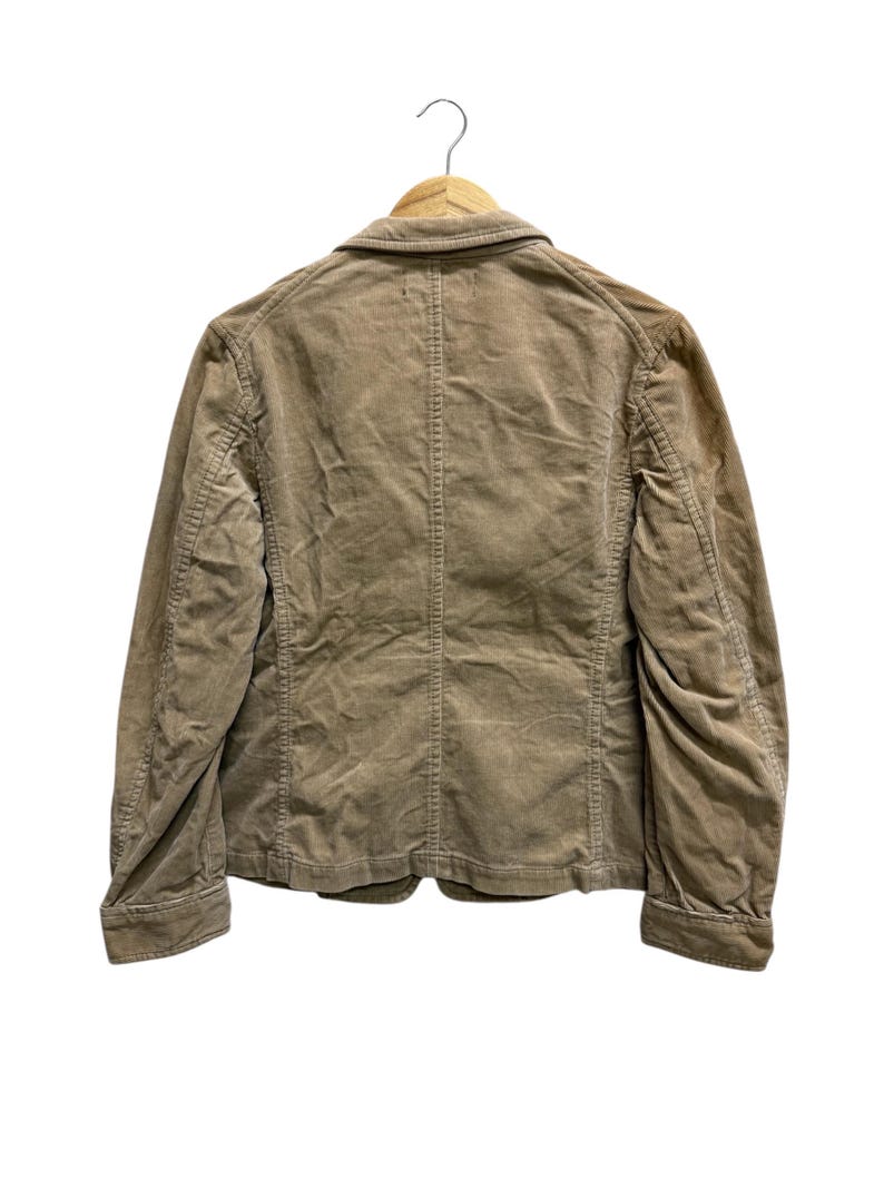 Puede incluir: Chaqueta de pana color beige, colgada de una percha de madera. La chaqueta presenta cierre de botones, cuello cl&aacute;sico y mangas largas. La vista trasera revela costuras verticales y un dise&ntilde;o estructurado, ideal para uso diario.