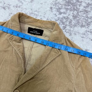 Puede incluir: Blazer de pana color beige con cierre de un solo bot&oacute;n. La etiqueta dice "tricot COMME des GAR&Ccedil;ONS". El blazer se mide con una cinta m&eacute;trica azul, aproximadamente 45 cm.