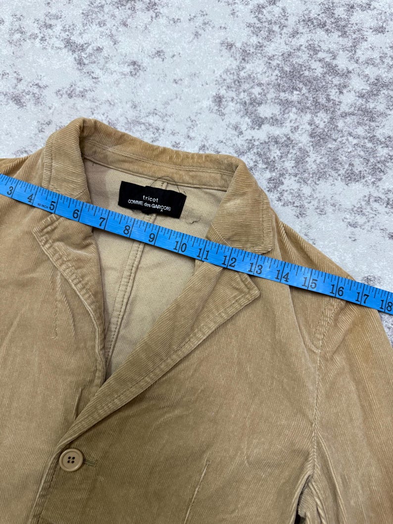 Puede incluir: Blazer de pana color beige con cierre de un solo bot&oacute;n. La etiqueta dice "tricot COMME des GAR&Ccedil;ONS". El blazer se mide con una cinta m&eacute;trica azul, aproximadamente 45 cm.