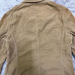 Puede incluir: Un blazer de pana color marr&oacute;n claro con cuello cl&aacute;sico y cierre de botones. La chaqueta presenta costillas de pana verticales y un corte entallado. La parte trasera del blazer es visible, mostrando las costuras y los detalles de construcci&oacute;n.