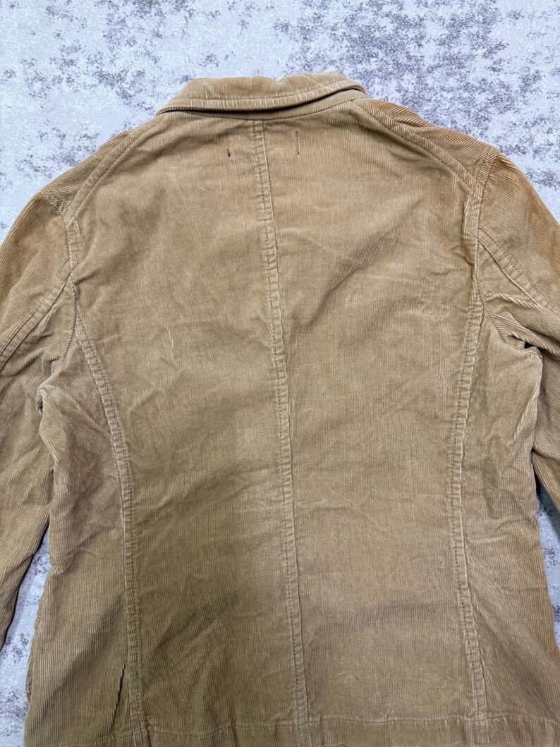 Puede incluir: Un blazer de pana color marr&oacute;n claro con cuello cl&aacute;sico y cierre de botones. La chaqueta presenta costillas de pana verticales y un corte entallado. La parte trasera del blazer es visible, mostrando las costuras y los detalles de construcci&oacute;n.