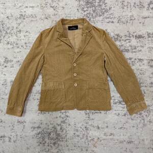 Puede incluir: Una blazer de pana color beige con solapa de muesca, cierre de tres botones y dos bolsillos delanteros. La chaqueta tiene mangas largas y un corte entallado. La etiqueta interior es visible. La blazer est&aacute; hecha de una tela suave y texturizada.