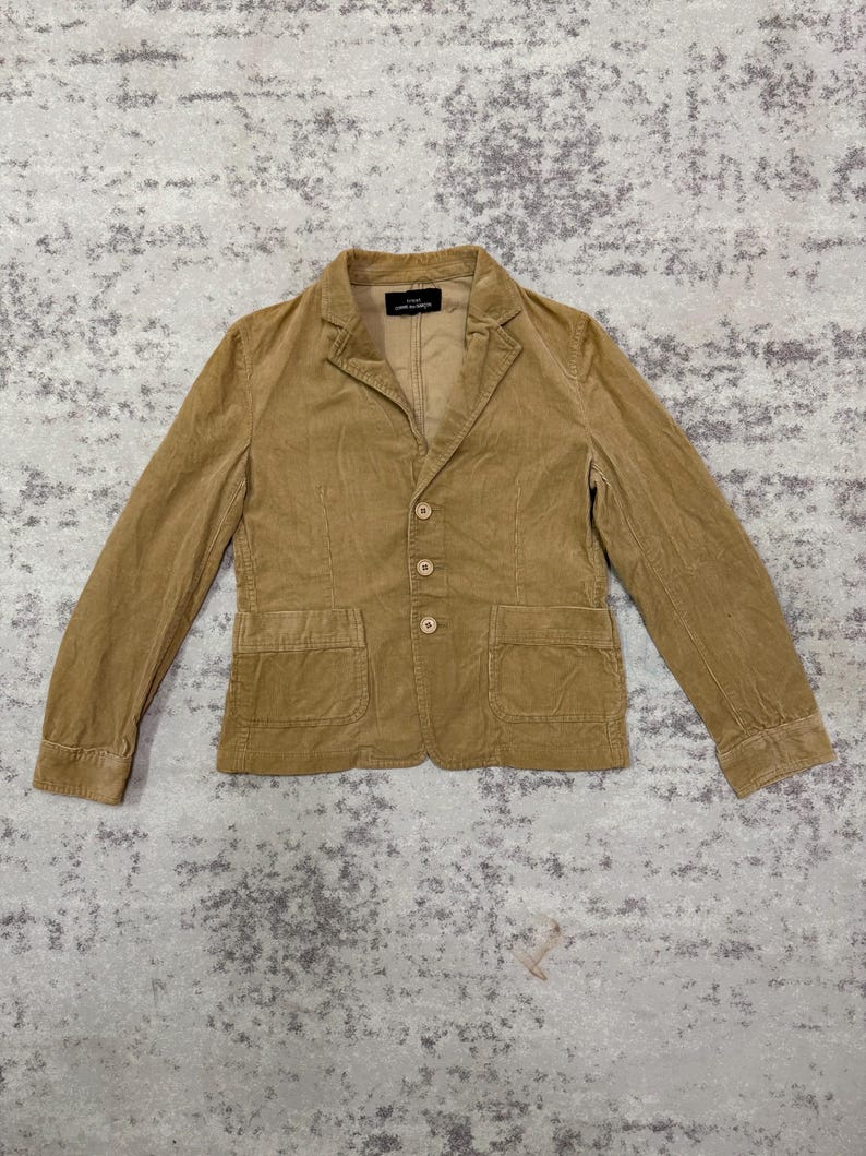 Puede incluir: Una blazer de pana color beige con solapa de muesca, cierre de tres botones y dos bolsillos delanteros. La chaqueta tiene mangas largas y un corte entallado. La etiqueta interior es visible. La blazer est&aacute; hecha de una tela suave y texturizada.