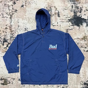 Puede incluir: Una chaqueta azul marino con capucha, cierre de presión y logotipo de Bud Sports en la parte delantera. La chaqueta tiene dos bolsillos en la parte delantera y una capucha con cordón.