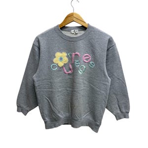 May include: A gray sweatshirt with the word "courage" embroidered in pastel colors. The word is stylized with a flower replacing the "o". The sweatshirt is hanging on a wooden hanger.