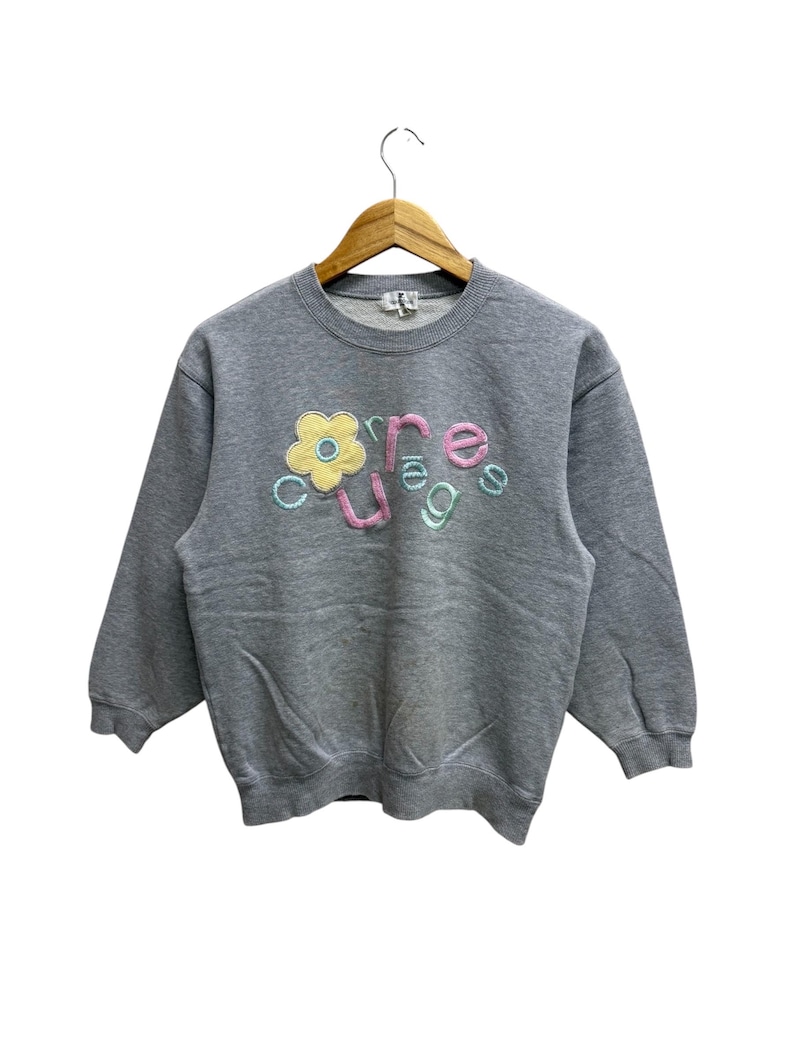 May include: A gray sweatshirt with the word "courage" embroidered in pastel colors. The word is stylized with a flower replacing the "o". The sweatshirt is hanging on a wooden hanger.