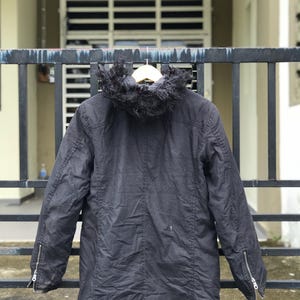 May include: A black hooded jacket featuring a faux fur trim on the hood, long sleeves with silver-toned metal zippers at the cuffs, and a central vertical seam down the back. The jacket is displayed on a wooden hanger against a dark metal gate.