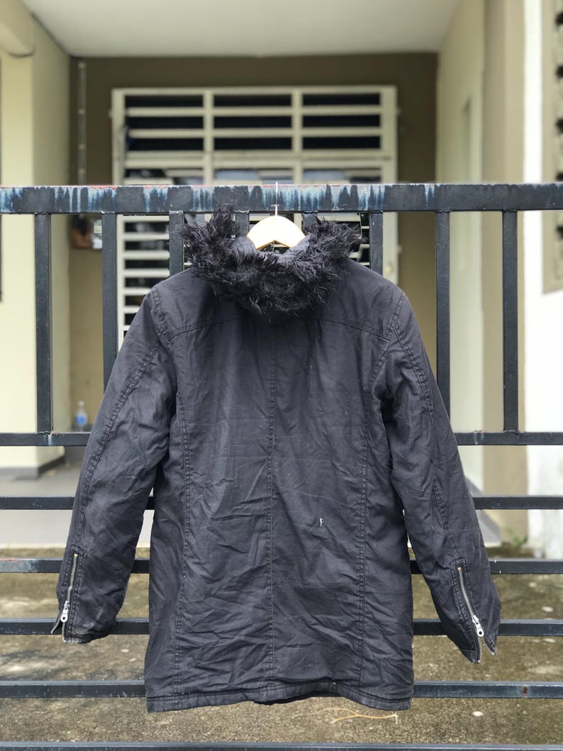 May include: A black hooded jacket featuring a faux fur trim on the hood, long sleeves with silver-toned metal zippers at the cuffs, and a central vertical seam down the back. The jacket is displayed on a wooden hanger against a dark metal gate.