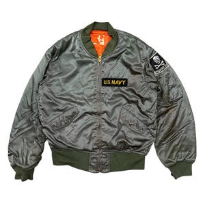 May include: A vintage olive green bomber jacket with a contrasting orange lining. Features a "U.S. NAVY" patch and a skull and crossbones patch. The jacket has a zip closure, two front pockets, and ribbed cuffs and hem.