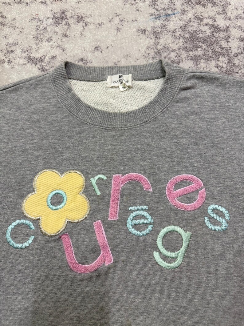 May include: A gray sweatshirt with a crew neck and embroidered floral and text design. The design features a yellow flower with a blue center and the word "cures" in pink, green, and blue lettering.