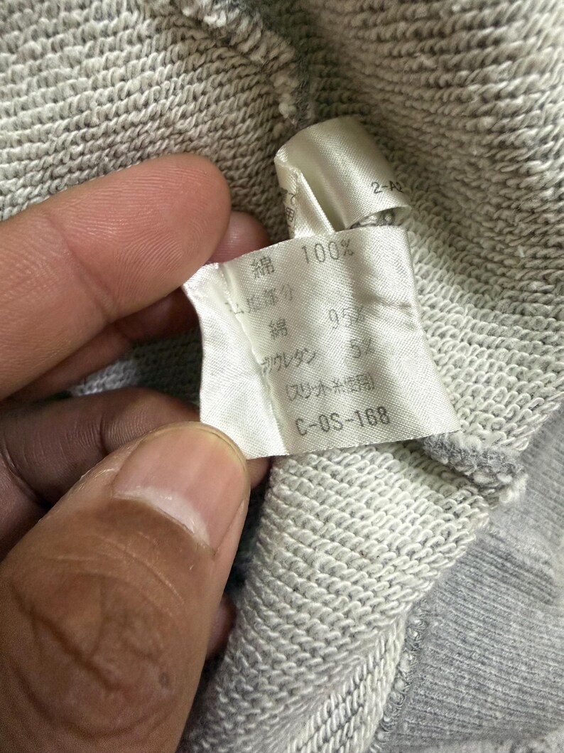May include: Close-up of a fabric tag with Japanese text and numbers. The tag is white with black text, showing fabric composition: 100% cotton, 95% cotton, and 5% polyurethane. The tag is attached to a gray fabric.