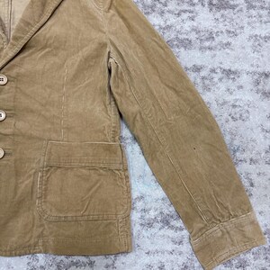 Puede incluir: Una blazer de pana color beige con solapa de muesca, tres botones y un bolsillo de parche. La chaqueta tiene un dise&ntilde;o cl&aacute;sico y un aspecto texturizado. La manga es visible y la tela tiene estr&iacute;as verticales.