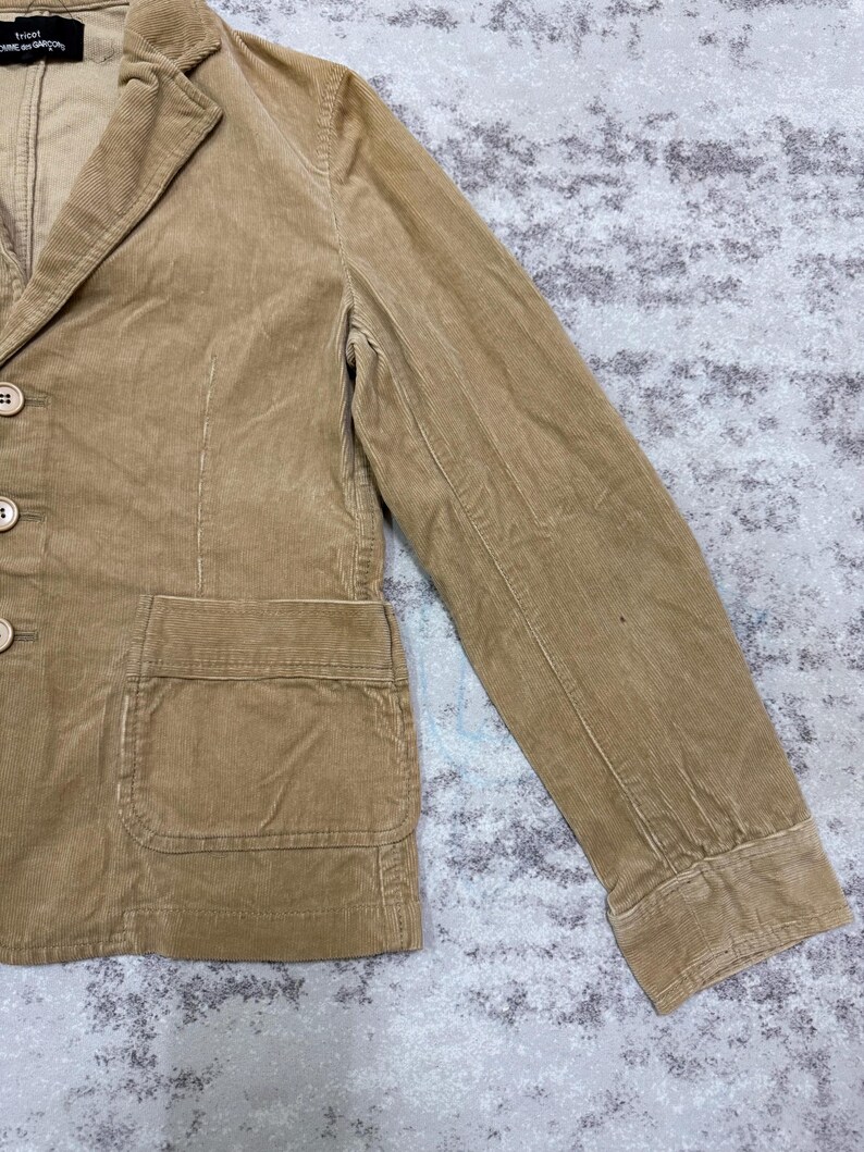 Puede incluir: Una blazer de pana color beige con solapa de muesca, tres botones y un bolsillo de parche. La chaqueta tiene un dise&ntilde;o cl&aacute;sico y un aspecto texturizado. La manga es visible y la tela tiene estr&iacute;as verticales.