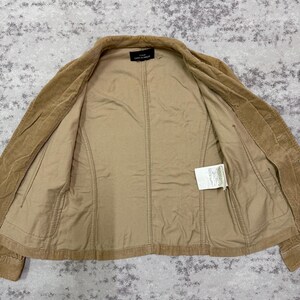 Puede incluir: Chaqueta de pana color canela con cierre de botones y corte entallado. La chaqueta tiene un forro interior marr&oacute;n claro y una peque&ntilde;a etiqueta. La chaqueta est&aacute; tendida sobre una superficie texturizada.