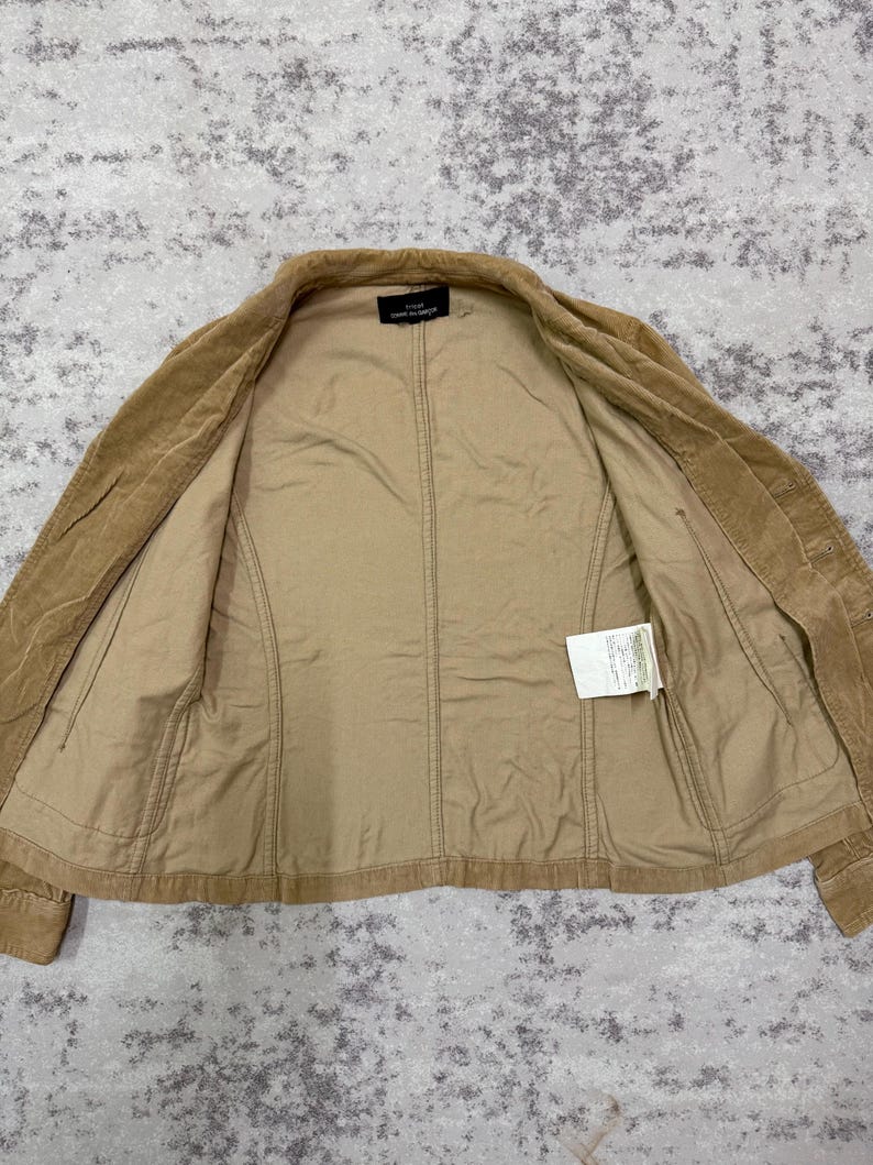 Puede incluir: Chaqueta de pana color canela con cierre de botones y corte entallado. La chaqueta tiene un forro interior marr&oacute;n claro y una peque&ntilde;a etiqueta. La chaqueta est&aacute; tendida sobre una superficie texturizada.