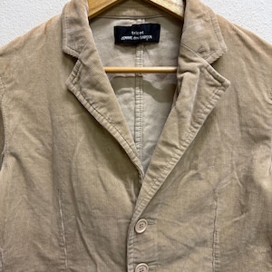 Puede incluir: Una blazer de pana color beige con solapa de muesca, cierre de tres botones y dos bolsillos delanteros. La blazer est&aacute; colgada en una percha de madera. La etiqueta dice "tricot COMME des GAR&Ccedil;ONS".