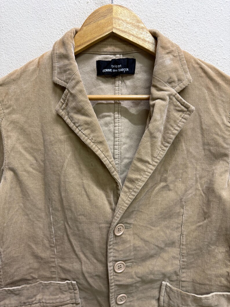 Puede incluir: Una blazer de pana color beige con solapa de muesca, cierre de tres botones y dos bolsillos delanteros. La blazer est&aacute; colgada en una percha de madera. La etiqueta dice "tricot COMME des GAR&Ccedil;ONS".