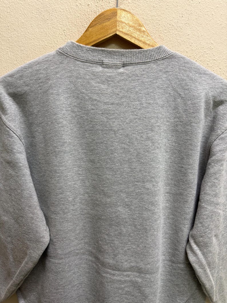 May include: A heather gray sweatshirt with a crew neck, hanging on a wooden hanger. The sweatshirt has a simple, minimalist design, with a focus on the soft, textured fabric. The sleeves are long and the overall style is casual.