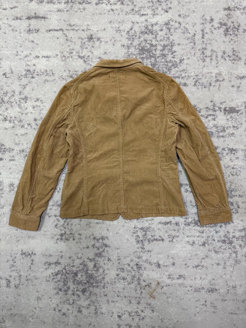 Puede incluir: Blazer de pana marr&oacute;n claro, visto desde atr&aacute;s. La chaqueta tiene un dise&ntilde;o cl&aacute;sico con cierre de bot&oacute;n y mangas largas. La tela de pana tiene una textura acanalada. El blazer es de color neutro.