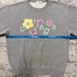 May include: A gray sweatshirt with the word "courage" embroidered in colorful letters and a flower design. The sweatshirt has a crew neck and long sleeves. The tape measure shows the sweatshirt is approximately 20 inches wide.