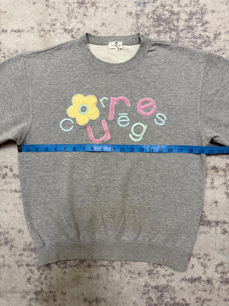 May include: A gray sweatshirt with the word "courage" embroidered in colorful letters and a flower design. The sweatshirt has a crew neck and long sleeves. The tape measure shows the sweatshirt is approximately 20 inches wide.