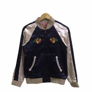 May include: Navy blue satin bomber jacket with embroidered rose details. The jacket has a zip-up front, side pockets, and contrasting silver sleeves and trim. A classic, stylish outerwear piece.