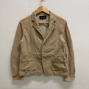 Puede incluir: Blazer de pana color beige con solapas de muesca, tres botones y dos bolsillos delanteros. La chaqueta est&aacute; colgada de una percha de madera contra una pared blanca. La etiqueta dice "COMME des GAR&Ccedil;ONS".