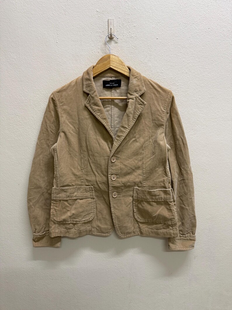 Puede incluir: Blazer de pana color beige con solapas de muesca, tres botones y dos bolsillos delanteros. La chaqueta est&aacute; colgada de una percha de madera contra una pared blanca. La etiqueta dice "COMME des GAR&Ccedil;ONS".