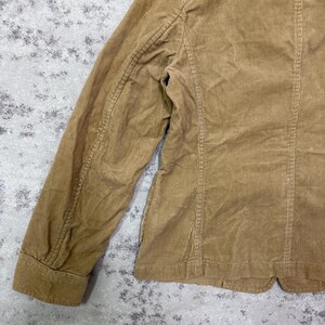 Puede incluir: Primer plano de una blazer de pana color beige. La chaqueta presenta un dise&ntilde;o cl&aacute;sico con solapa de muesca, cierre de bot&oacute;n y mangas largas. La tela de pana tiene una textura acanalada.