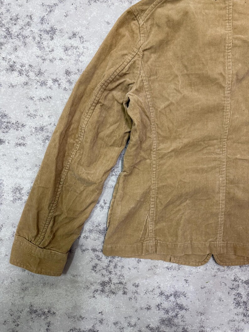 Puede incluir: Primer plano de una blazer de pana color beige. La chaqueta presenta un dise&ntilde;o cl&aacute;sico con solapa de muesca, cierre de bot&oacute;n y mangas largas. La tela de pana tiene una textura acanalada.