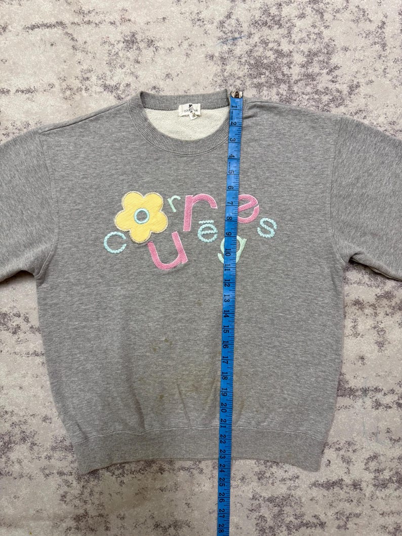 May include: A gray sweatshirt with a crew neck and long sleeves. The front features the word "curious" in a playful font, with a flower graphic. The sweatshirt is measured with a blue tape measure.