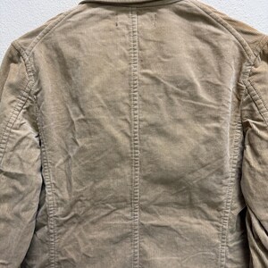 Puede incluir: Primer plano de una blazer de pana color beige. Se ve la parte trasera de la chaqueta, mostrando el cuello, las costuras y la textura vertical de la pana. La chaqueta est&aacute; colgada en una percha de madera.