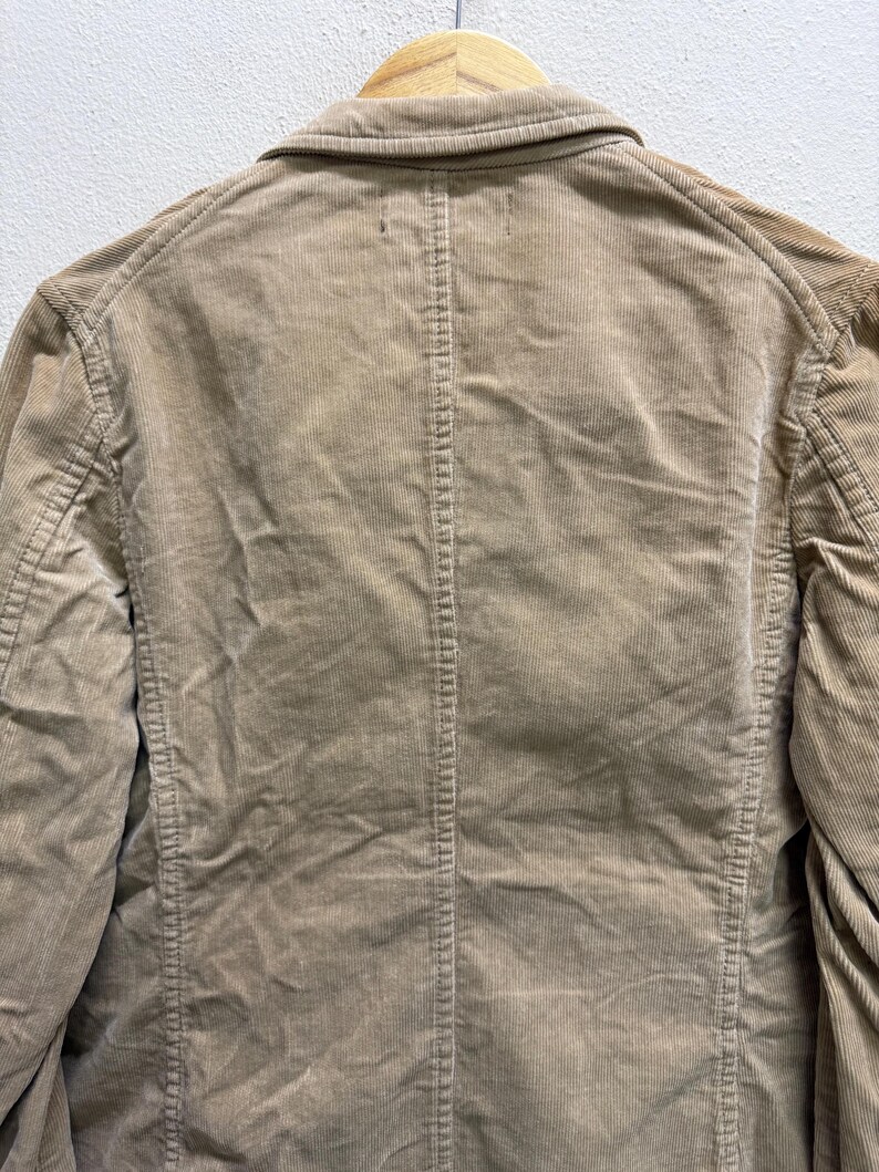 Puede incluir: Primer plano de una blazer de pana color beige. Se ve la parte trasera de la chaqueta, mostrando el cuello, las costuras y la textura vertical de la pana. La chaqueta est&aacute; colgada en una percha de madera.