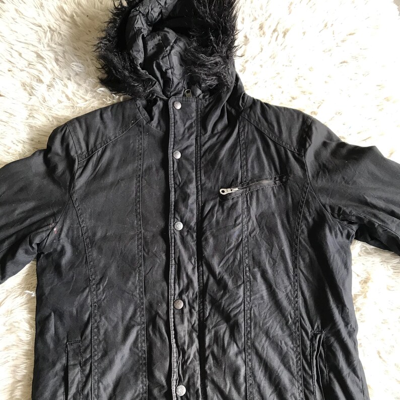 May include: A black hooded jacket featuring a faux fur trim on the hood, a front button placket with metallic snap closures, long sleeves, and a single zippered chest pocket. The jacket is constructed with vertical seam detailing along the front.