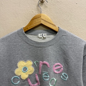 May include: Gray crew neck sweatshirt with a floral design and the word "Cure" embroidered in pink, green, and blue. The flower is yellow with a blue center. The sweatshirt is hanging on a wooden hanger.