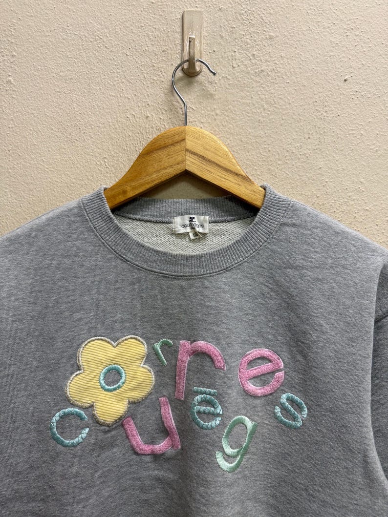 May include: Gray crew neck sweatshirt with a floral design and the word "Cure" embroidered in pink, green, and blue. The flower is yellow with a blue center. The sweatshirt is hanging on a wooden hanger.