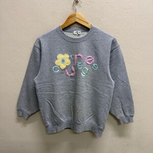 May include: A gray sweatshirt with the word "courage" embroidered in pastel colors. The word is arranged with a flower graphic. The sweatshirt is hanging on a wooden hanger.