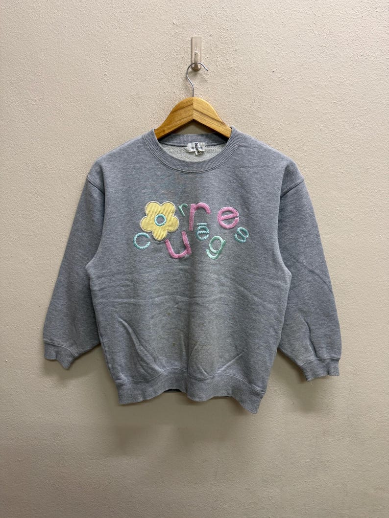 May include: A gray sweatshirt with the word "courage" embroidered in pastel colors. The word is arranged with a flower graphic. The sweatshirt is hanging on a wooden hanger.