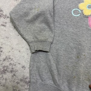 May include: A gray sweatshirt with a floral design on the front. The flower is yellow with a blue center and a pink letter "U" below it. The sweatshirt has a ribbed hem and cuffs.