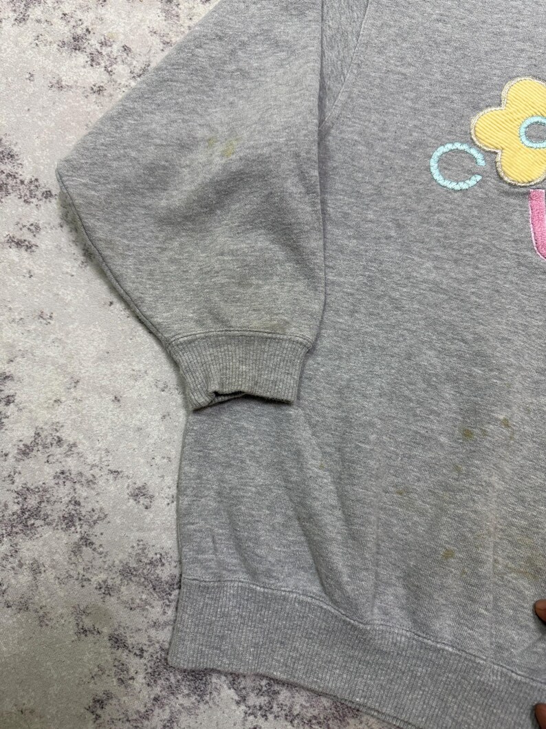 May include: A gray sweatshirt with a floral design on the front. The flower is yellow with a blue center and a pink letter "U" below it. The sweatshirt has a ribbed hem and cuffs.