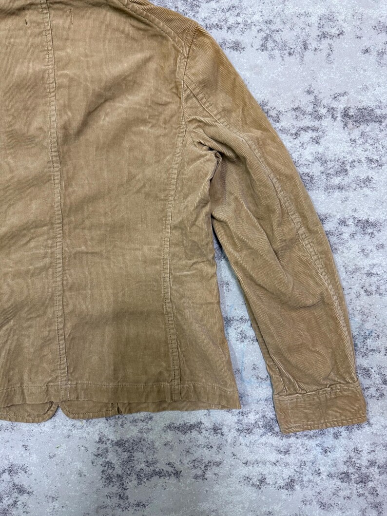 Puede incluir: Un blazer de pana color beige con un dise&ntilde;o cl&aacute;sico. La chaqueta presenta solapas con muescas, cierre de dos botones y un corte entallado. La tela de pana tiene una textura acanalada. Se ve la manga.