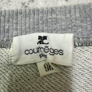 May include: Close-up of a gray sweatshirt with a white tag. The tag features the brand name "courr&egrave;ges paris" in black text and a small logo. A smaller tag with the number "9H" is attached.