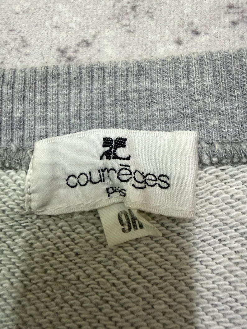 May include: Close-up of a gray sweatshirt with a white tag. The tag features the brand name "courr&egrave;ges paris" in black text and a small logo. A smaller tag with the number "9H" is attached.