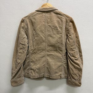 Puede incluir: Chaqueta de pana color beige claro con cierre de botones, colgada de una percha de madera. La chaqueta tiene un dise&ntilde;o cl&aacute;sico con cuello con solapas y dos bolsillos delanteros. La tela de pana tiene una textura acanalada.