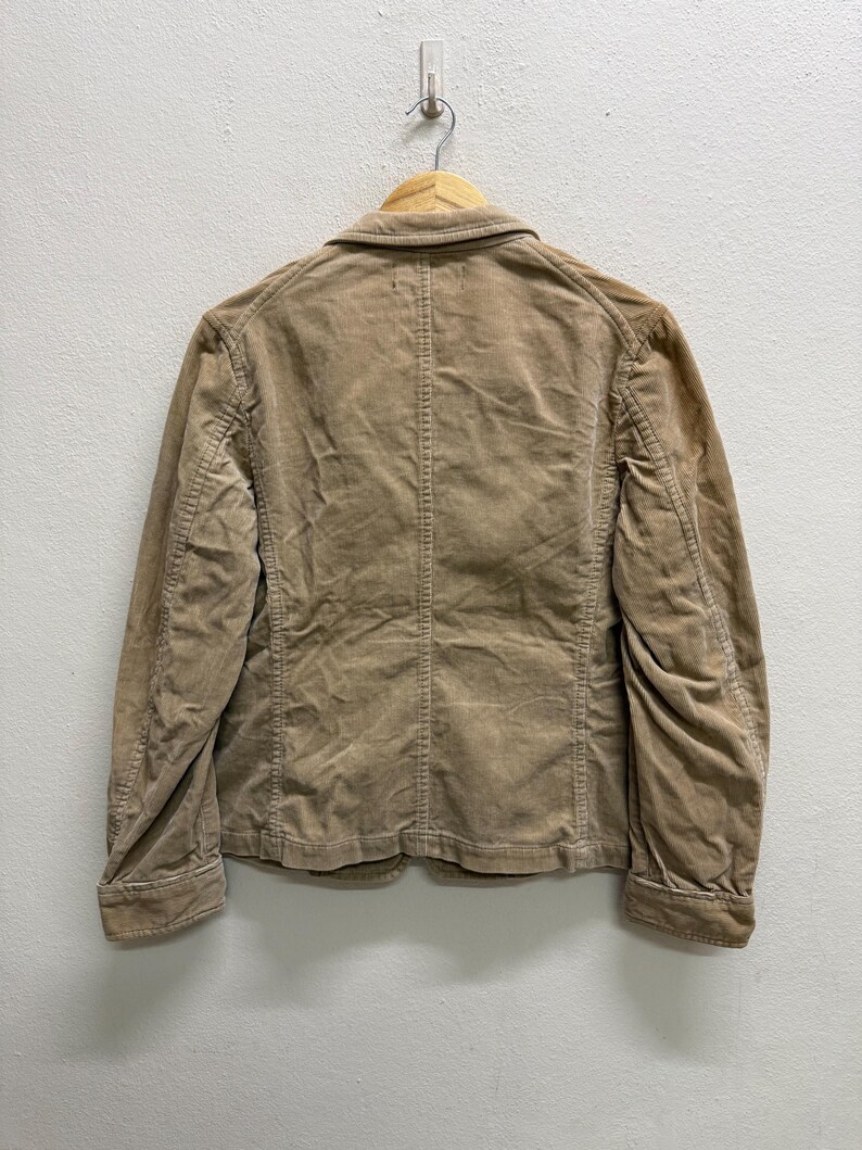 Puede incluir: Chaqueta de pana color beige claro con cierre de botones, colgada de una percha de madera. La chaqueta tiene un dise&ntilde;o cl&aacute;sico con cuello con solapas y dos bolsillos delanteros. La tela de pana tiene una textura acanalada.