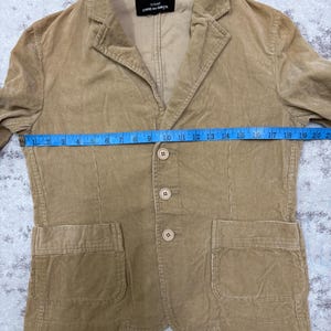 Puede incluir: Blazer de pana color beige con solapas de muesca, cierre de tres botones y dos bolsillos delanteros. El blazer tiene una etiqueta que dice "tricot COMME des GAR&Ccedil;ONS". El blazer mide aproximadamente 48 cm en el pecho.