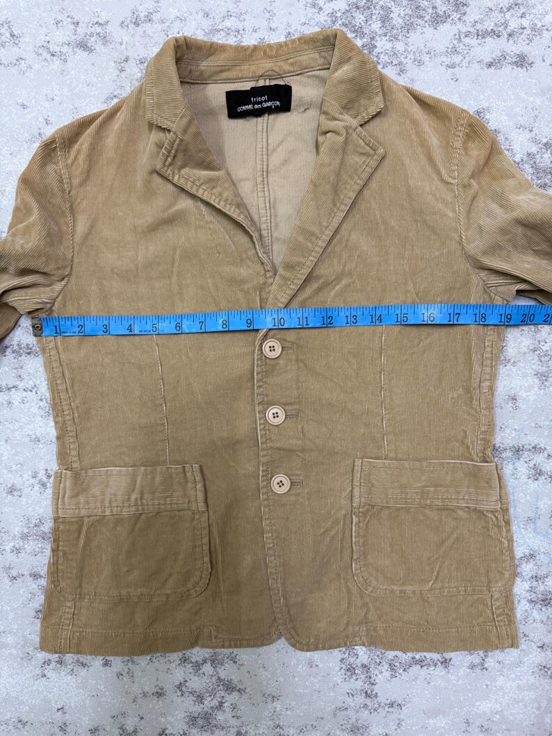 Puede incluir: Blazer de pana color beige con solapas de muesca, cierre de tres botones y dos bolsillos delanteros. El blazer tiene una etiqueta que dice "tricot COMME des GAR&Ccedil;ONS". El blazer mide aproximadamente 48 cm en el pecho.