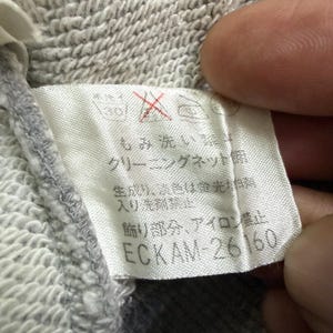 May include: Close-up of a clothing tag with Japanese text and the number ECKAM-26160. The tag is white with black text and a red symbol. The fabric is a light gray, textured material.